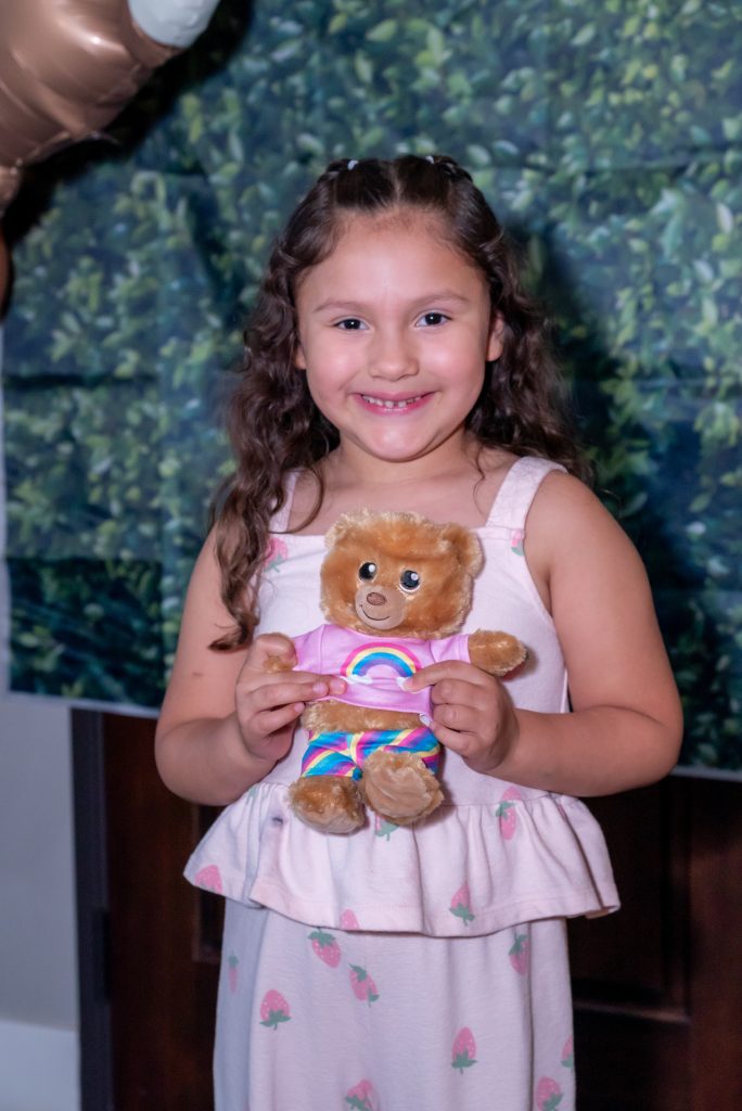 Smiling young girl with long brown hair wears a light pink dress with small designs and holds a brown teddy bear dressed in a rainbow shirt and shorts. A leafy green backdrop and balloons are behind her.