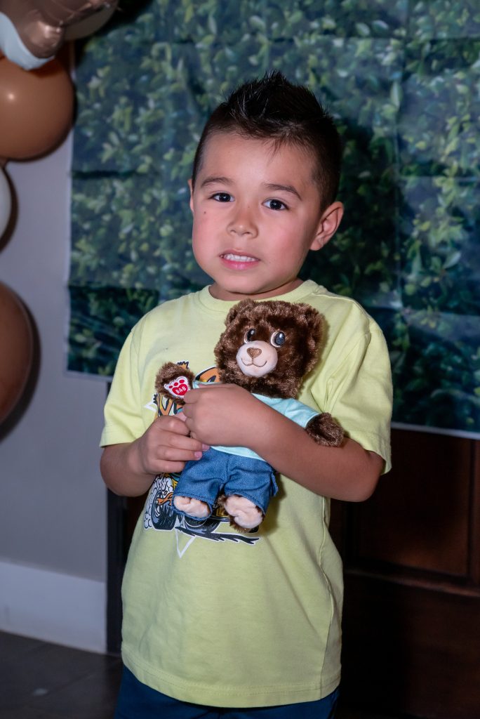 A young boy with short dark hair, wearing a yellow shirt, holds a brown teddy bear and smiles nervously. Green and brown balloons and a leafy backdrop are visible in the background.