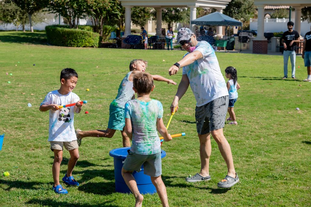 A man and three children in color-stained clothes have a water gun fight on a grassy park lawn.