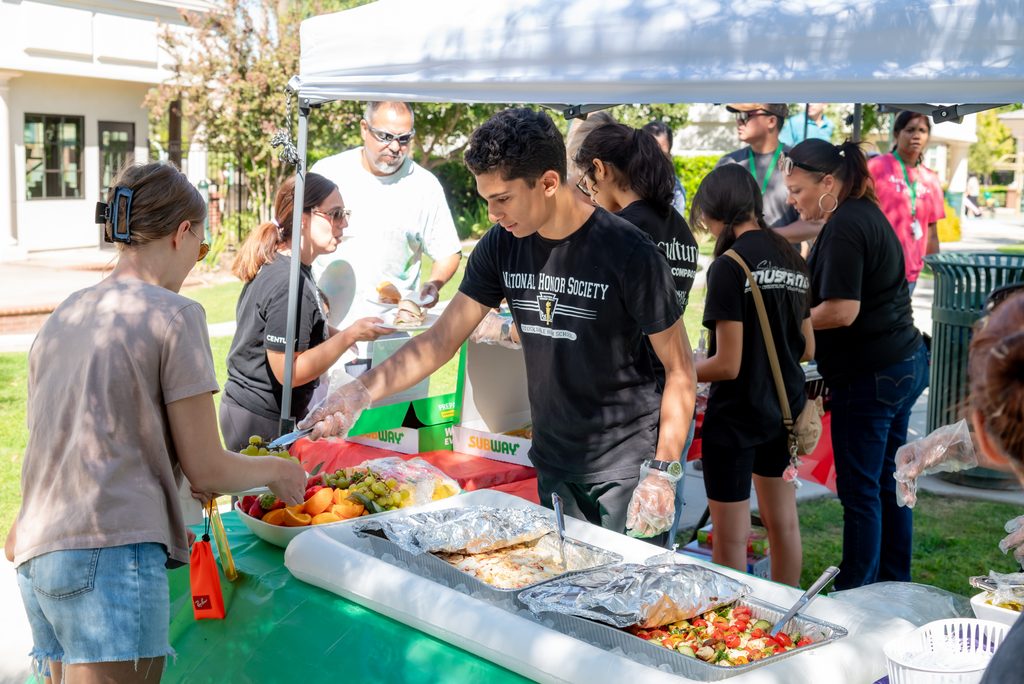 A National Honor Society volunteer wearing gloves serves food to a person at an outdoor community event.