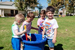 Four children in colorful, paint-splattered shirts stand around a blue barrel of water with spray toys, with one child in the foreground smiling at the camera.