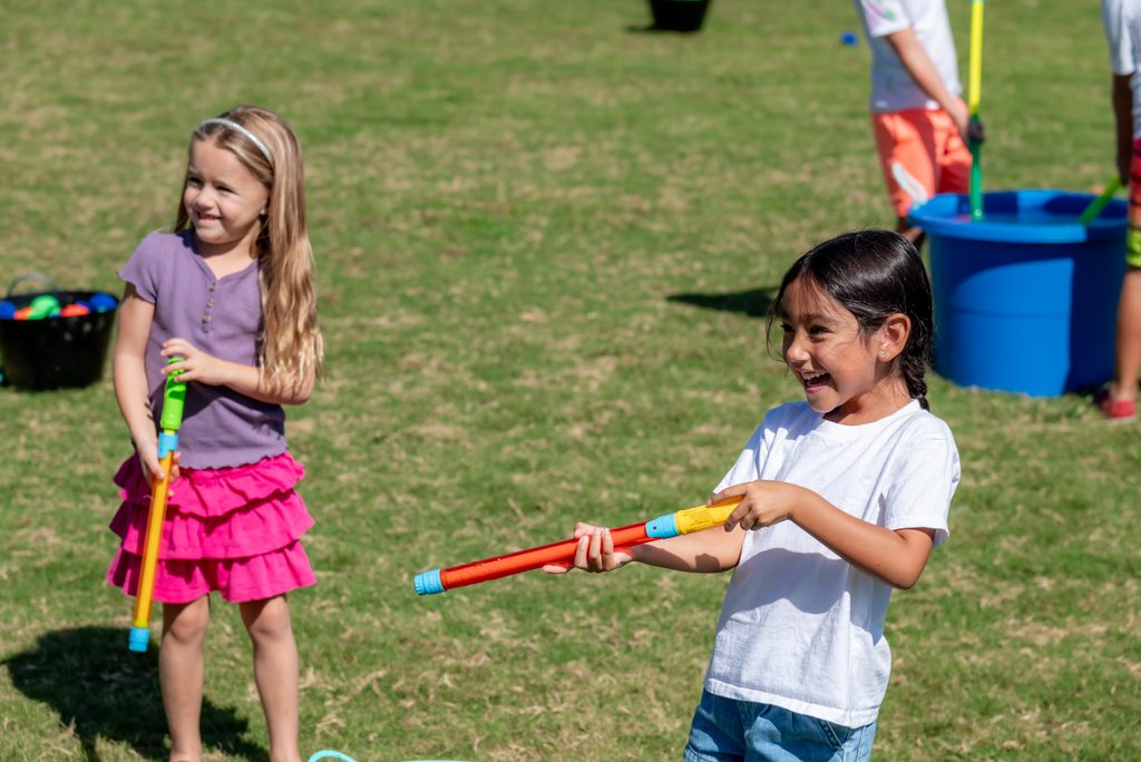 Two children playing with colorful foam water shooters on a grass field.