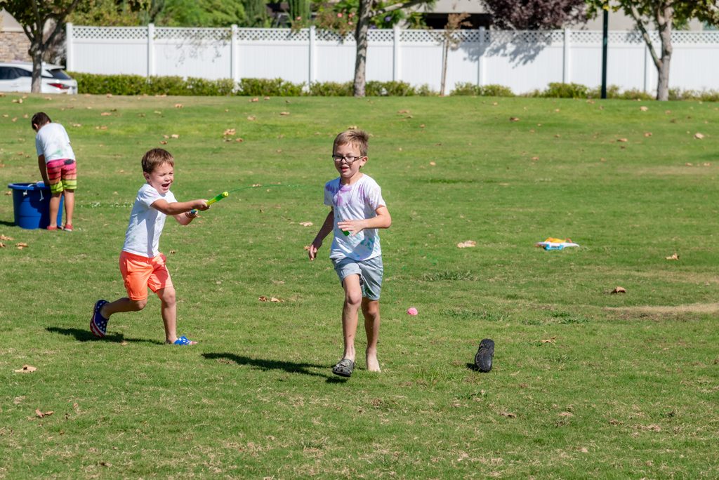 Two boys run across a grassy field while playing with a green squirt gun.