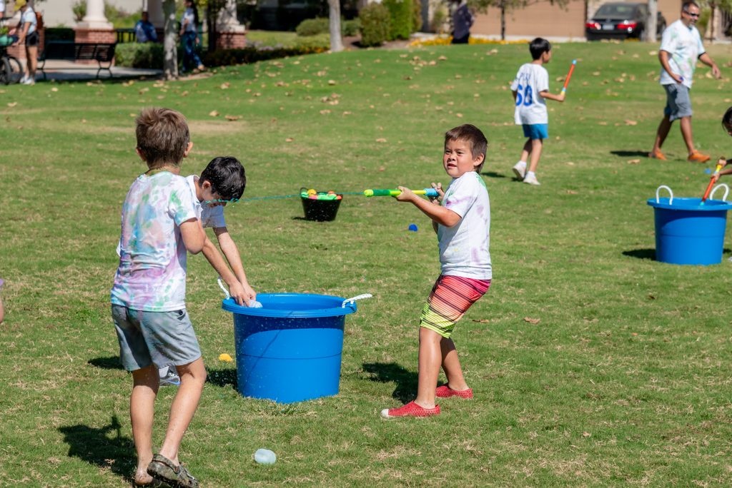 Children in tie-dye shirts have a water fight with squirt guns and buckets on a grassy field.