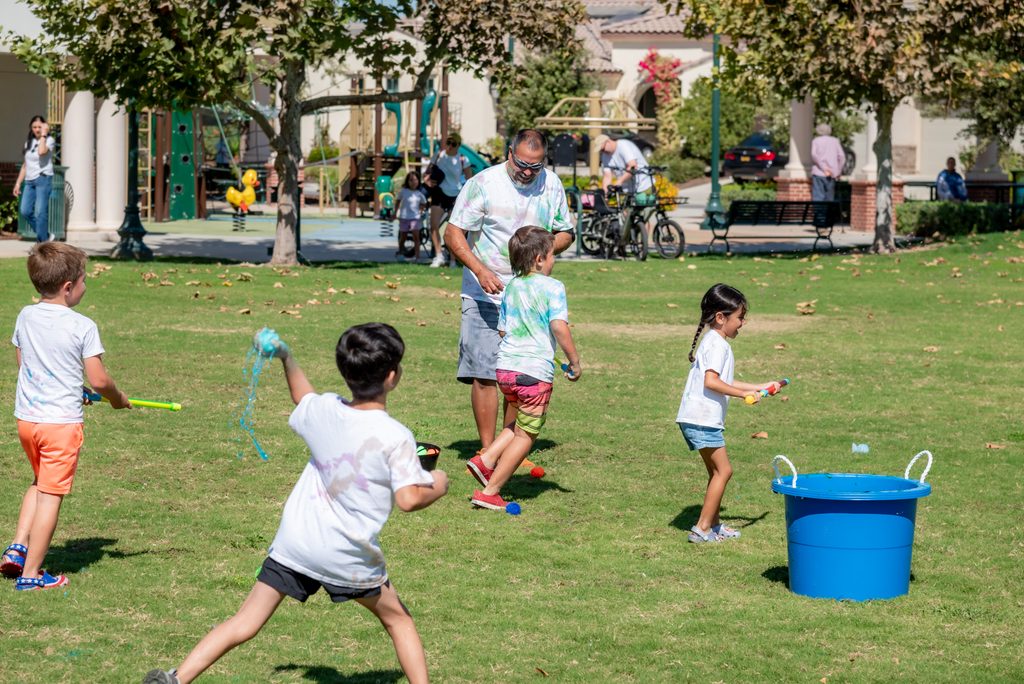 Children in tie-dye shirts play with water balloons on a park lawn near an adult and a large blue bucket.