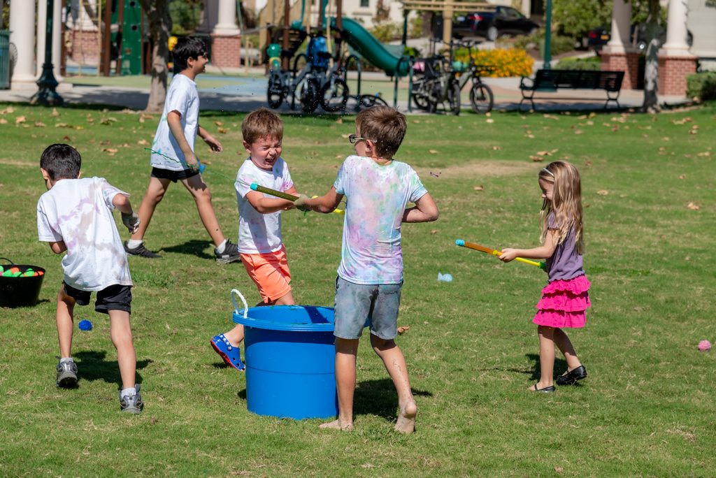Four children in tie-dye shirts play with water guns and a large blue bucket on a grassy park lawn.