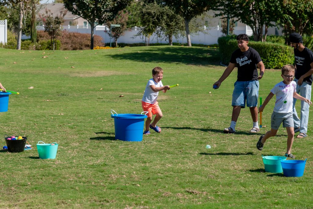 Three children and two adults participate in a water balloon game on a grassy field, using buckets to store balloons.