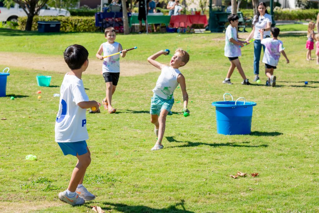 A boy in a white tank top throws a green water balloon at a friend during a grassy park event.