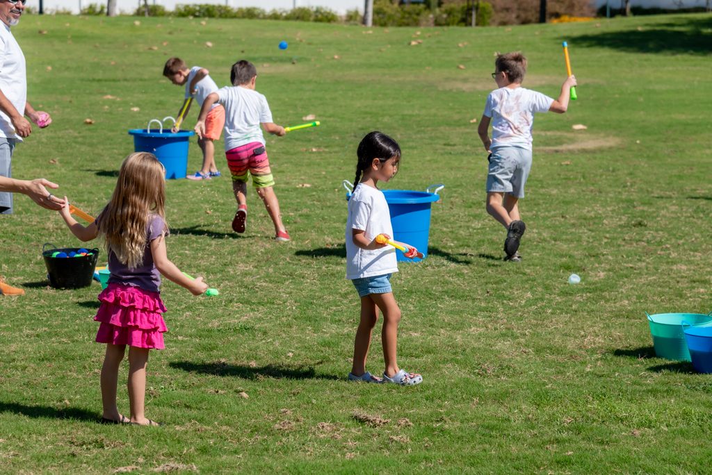 Children play with water blasters and plastic buckets on a grassy field.