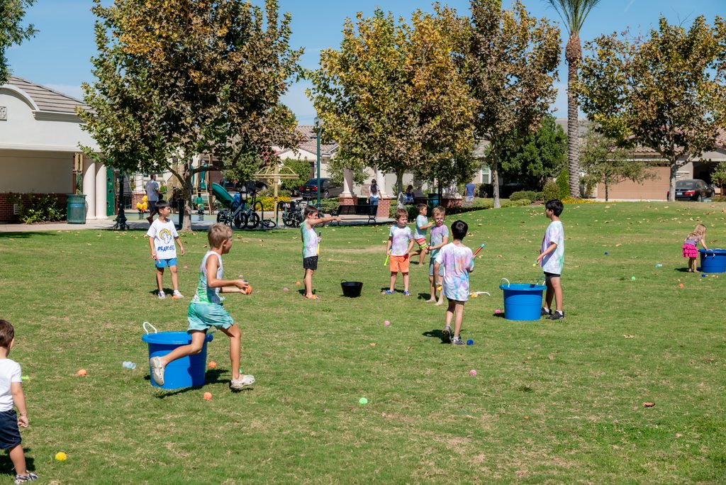 A group of children in a park participate in a water balloon game, holding balloons and running between blue plastic buckets.