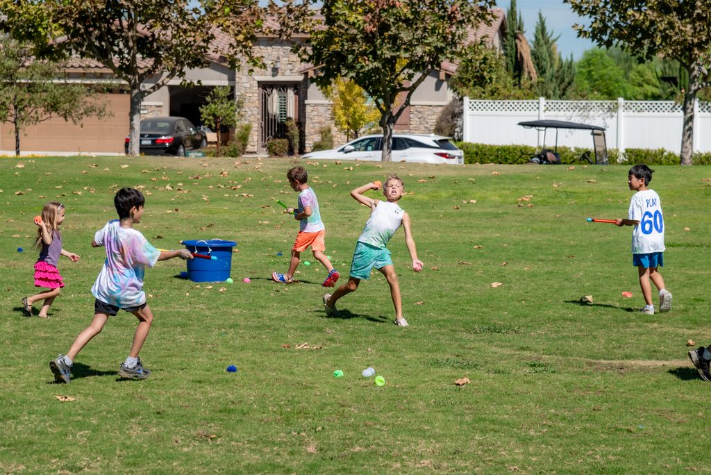 Five children playing with colorful water balloons and handheld launchers on a grassy park lawn.