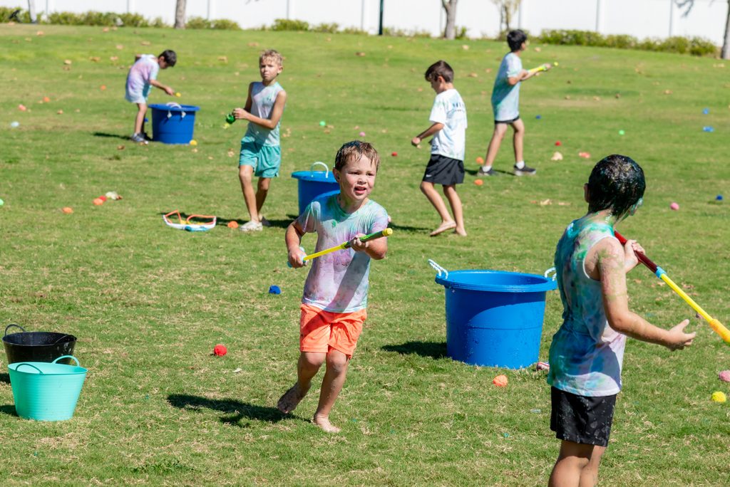 Children in white shirts covered in colorful powder play with water squirt guns and blue buckets on a grassy field.