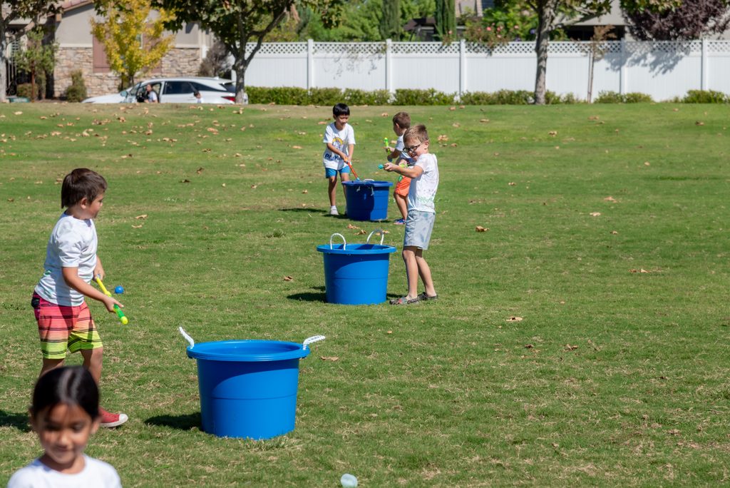 Children participate in a water balloon game on a grassy field, each aiming to toss balloons into their own blue bucket.