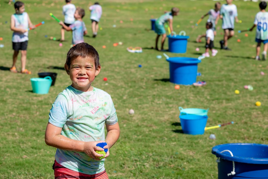 A smiling child holding a blue and green sponge stands on a field with other children playing near blue water buckets.