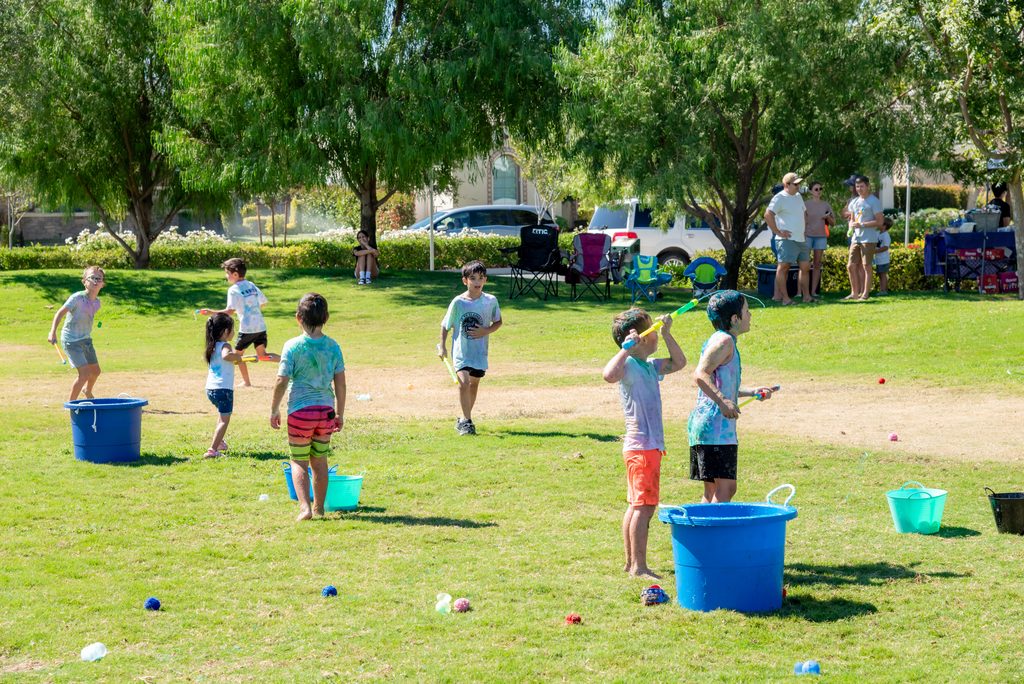 Children play with water guns and water balloons on a sunny park lawn, with blue buckets scattered throughout the grass.