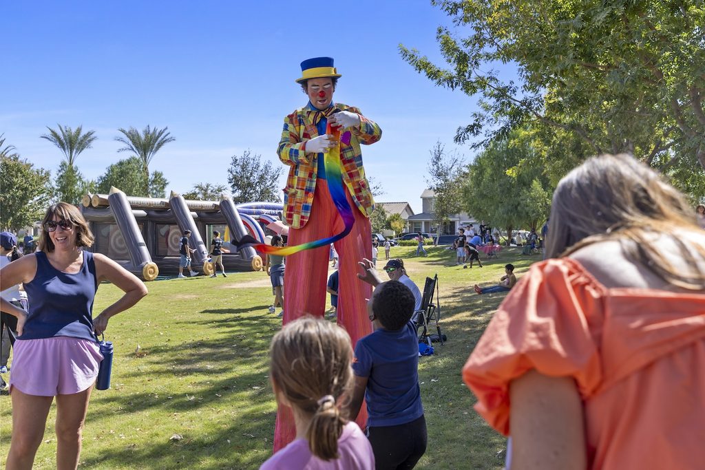 A stilt-walking clown in a colorful suit and top hat holds a long rainbow ribbon for children in a sunny park with an inflatable obstacle course in the background.