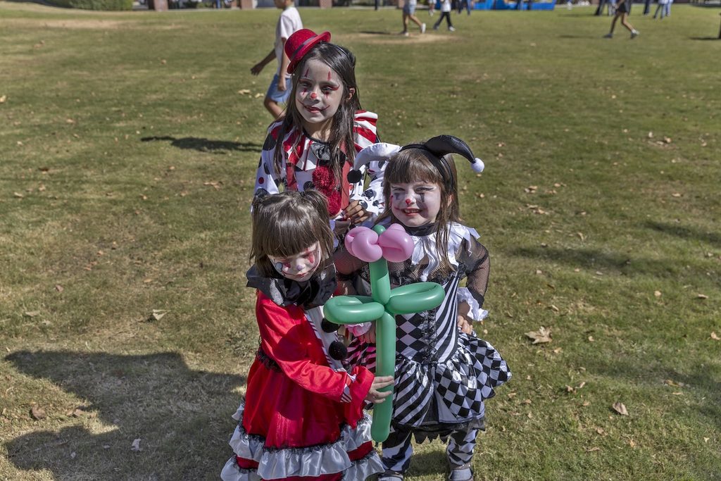 Three children in clown costumes and face paint standing on a grassy field, with one child holding a green and pink balloon flower.