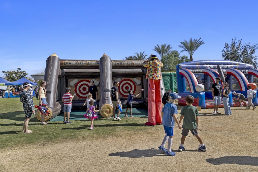 People at an outdoor festival playing at an inflatable lumberjack axe-throwing game and a sports-themed bounce house, with a stilt walker in a colorful suit standing near the axe-throwing station.