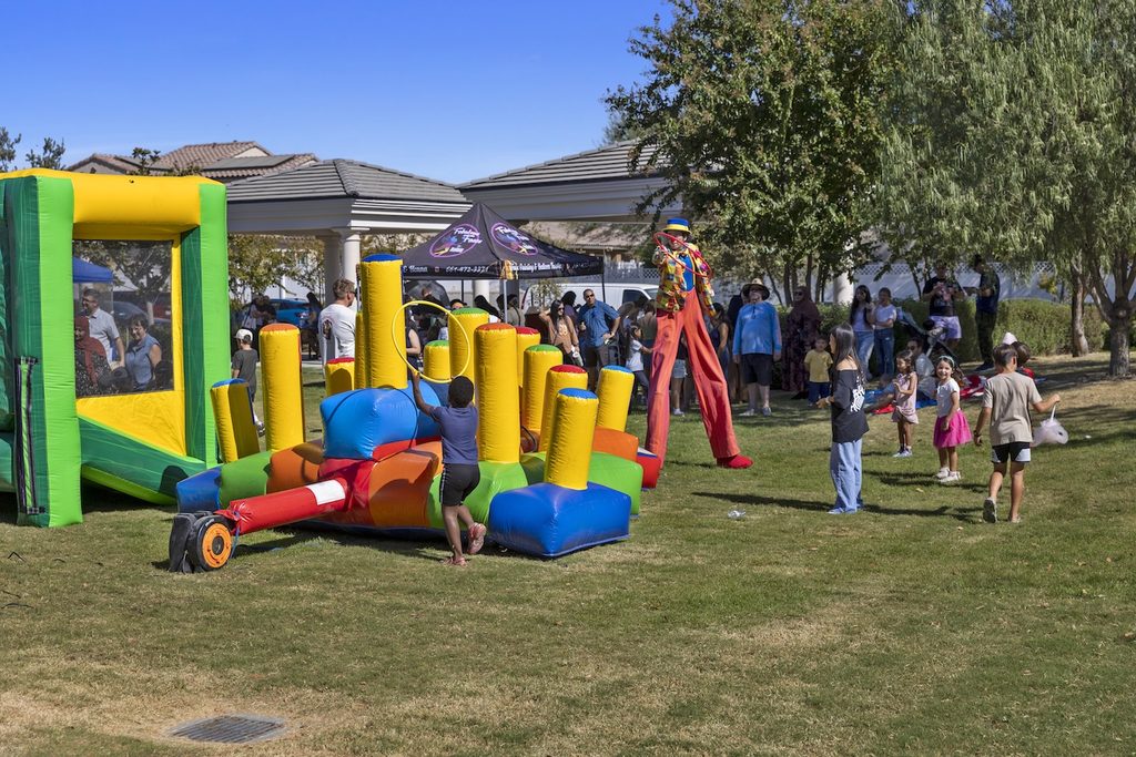 A stilt walker in a colorful clown outfit entertains children playing with a small inflatable obstacle course in a park.