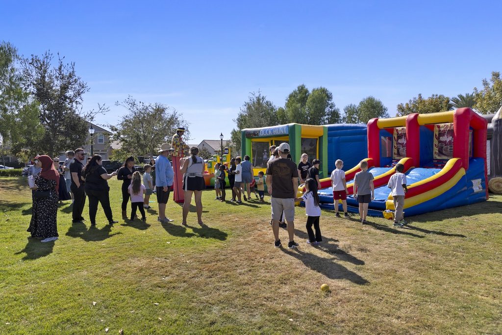 A group of adults and children gather in a park around several colorful inflatable bounce houses.