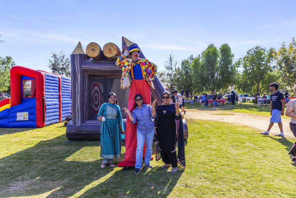 Three women posing for a photo in front of a stilt-walking clown at an outdoor park event with bounce houses.