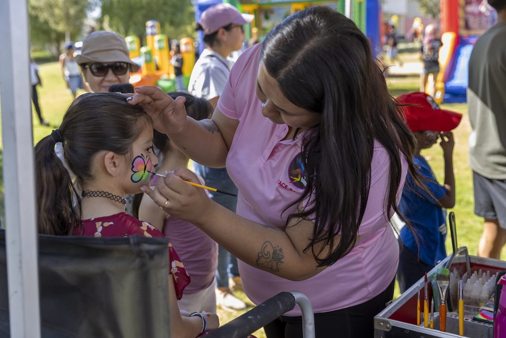 A face painter in a pink shirt applies a butterfly design to a young girl’s cheek at an outdoor event.