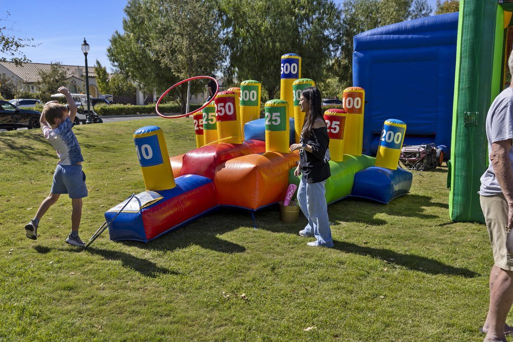 A boy throws a red ring at an inflatable ring-toss game with numbered targets, while a woman watches nearby on a grassy lawn.
