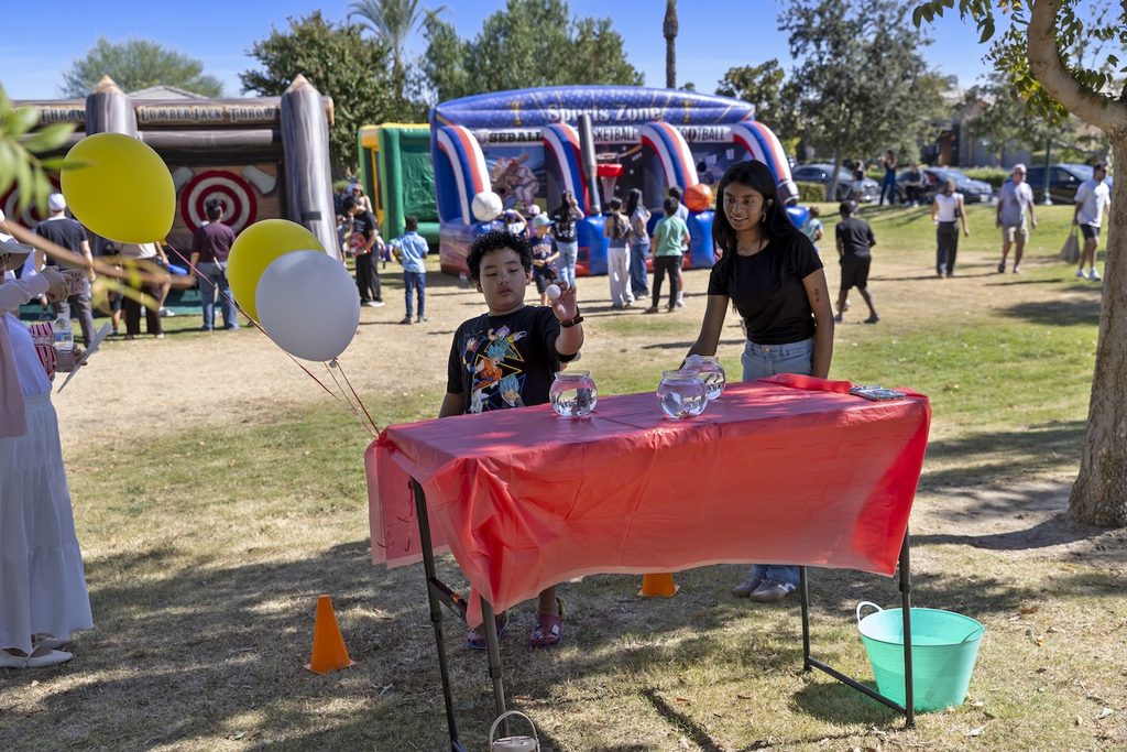 A boy and a girl stand at a table playing a goldfish toss game in a park with bounce houses in the background.