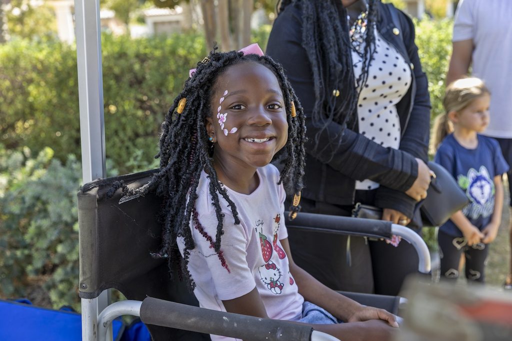 A young girl with dark locs and a flower-themed face painting smiles while sitting in a director’s chair.