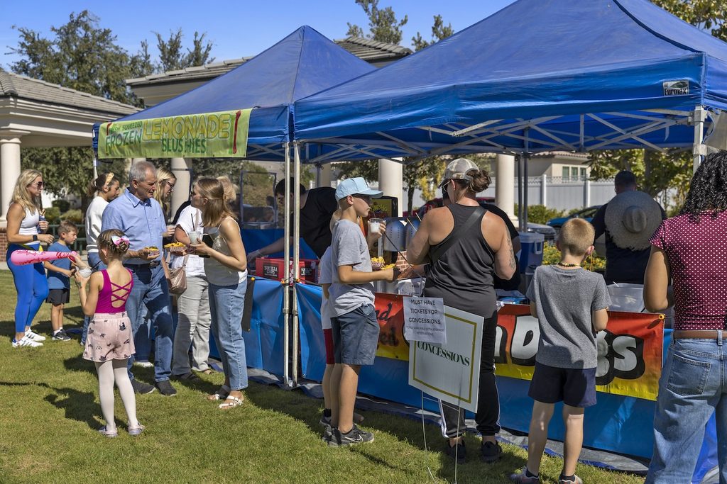 People at an outdoor food stand under blue tents, with signs for “Fresh Squeezed Frozen Lemonade Slushies” and “Concessions.”
