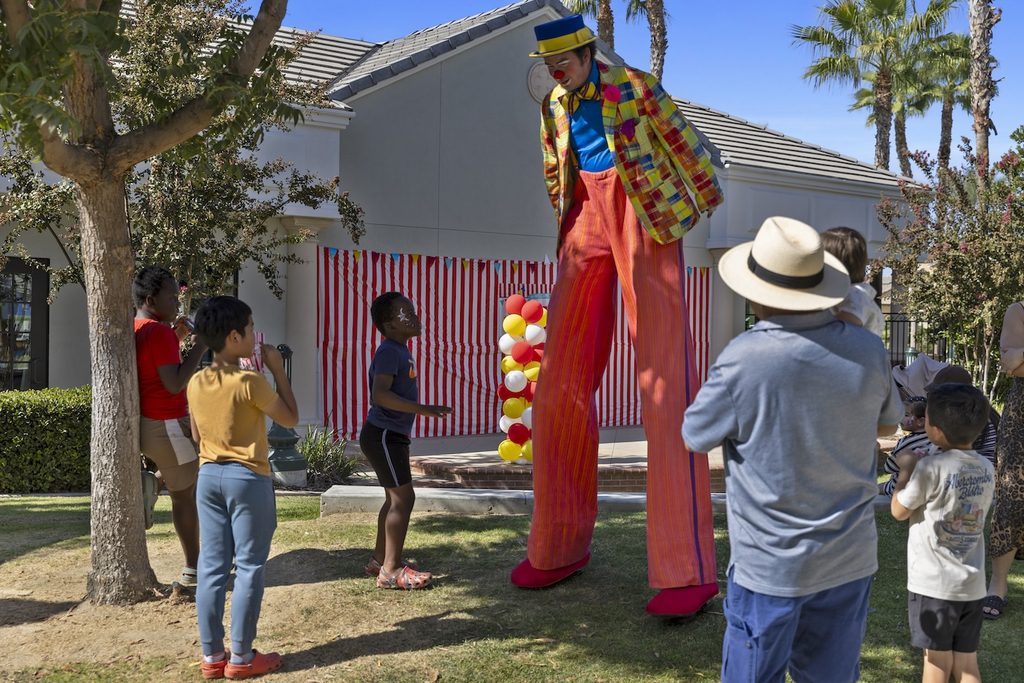 A stilt-walking clown in a colorful patchwork jacket and bright orange trousers entertains a group of children and adults outdoors.