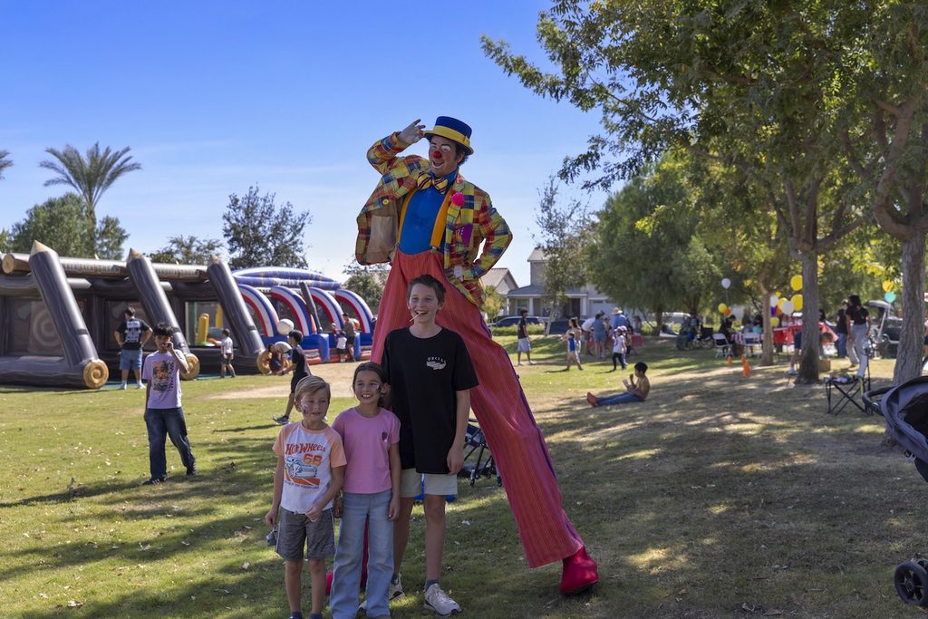 A stilt-walking clown in a colorful suit poses for a photo with three children in a park.